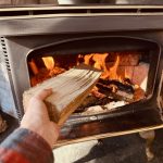 Hand adding a log to a burning fire inside a wood stove, providing warmth and comfort in a cozy indoor setting.