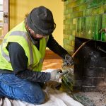 A person in a safety vest repairs a fireplace with green tiles.
