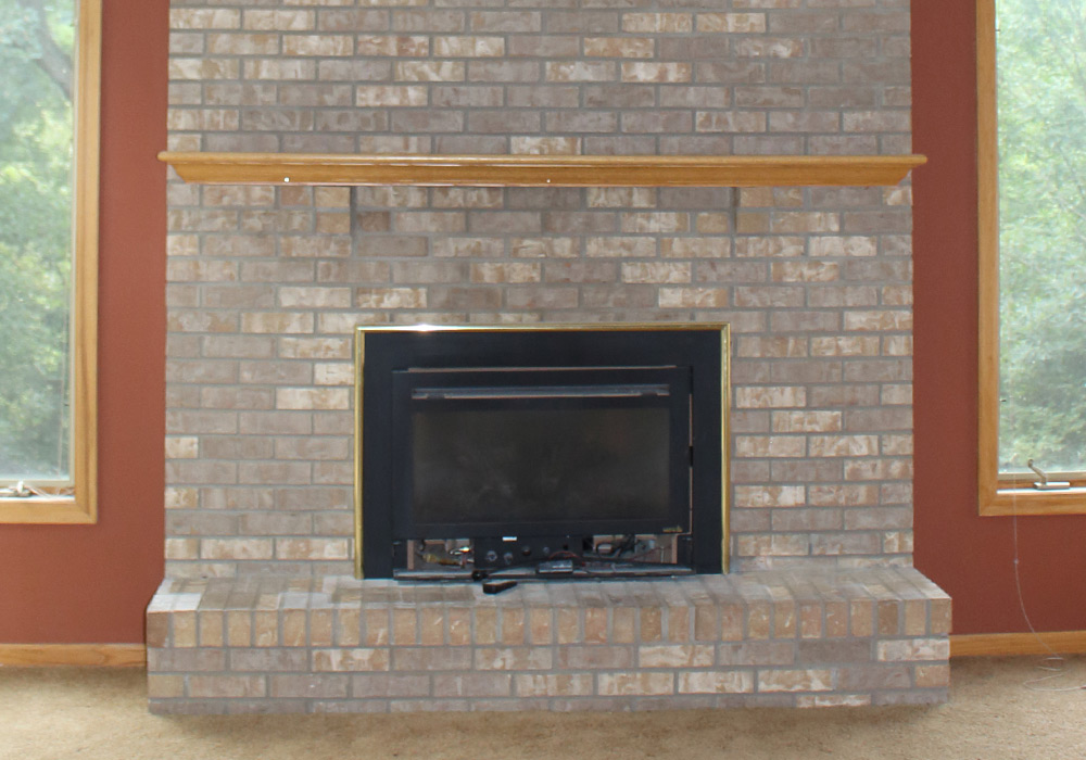 Brick fireplace with wooden mantel, flanked by windows, in a cozy living room setting.