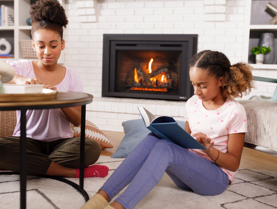 Two girls sitting by a fireplace, one reading a book, the other writing, in a cozy living room setting.