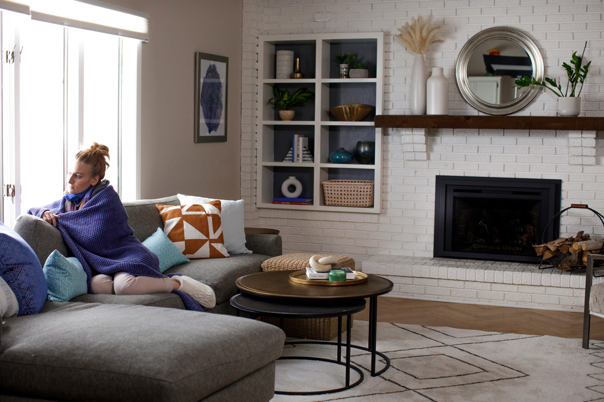 Woman wrapped in a purple blanket, sitting on a gray sofa in a cozy, modern living room.