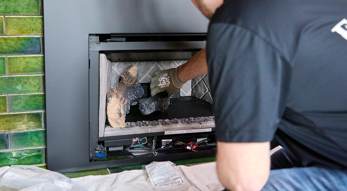 A person installing gas logs in a fireplace with green tile background.