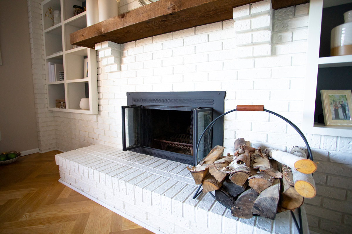White brick fireplace with wood mantel and log storage in a cozy living room setting.