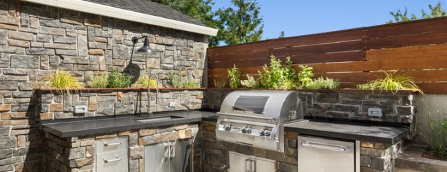 Outdoor kitchen with stone walls, stainless steel appliances, and a built-in grill under a clear blue sky.