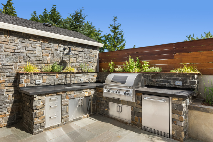 Outdoor kitchen with stone walls, stainless steel appliances, and a built-in grill under a clear blue sky.